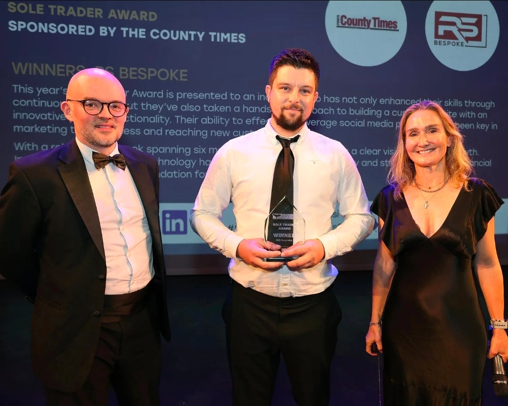 Three people standing together with an award, sponsored by The County Times and RS, against a backdrop with text and logos.